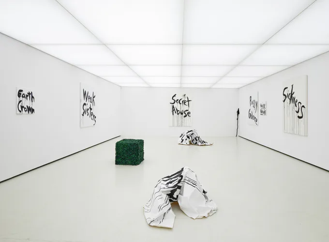 White gallery room with hand-drawn text on walls, green moss-covered cubic sculpture, crumpled white forms on floor, and overhead grid ceiling.