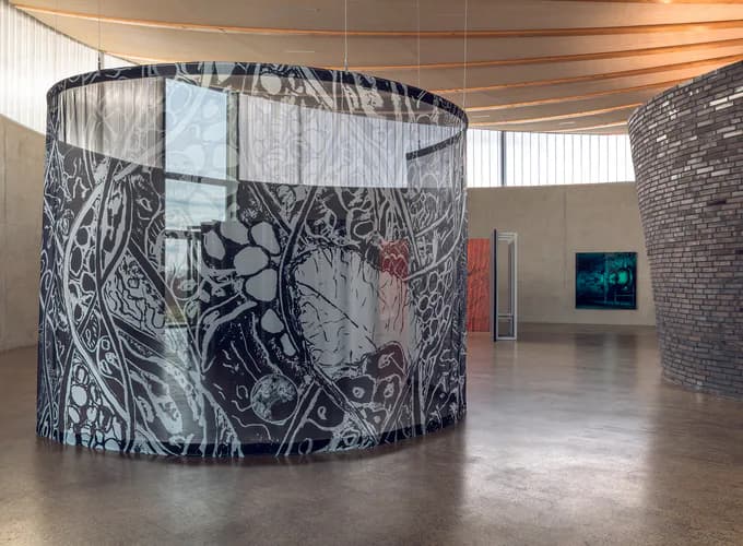 Gallery interior with large cylindrical installation wrapped in black and white intricate drawings, brick wall, and minimal furnishings under industrial ceiling.