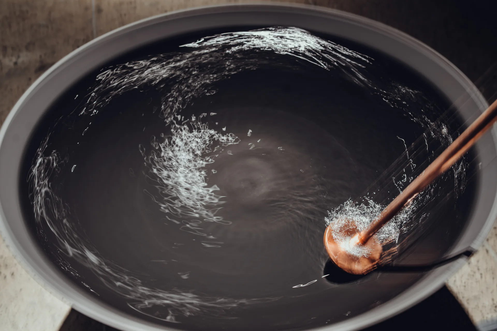 Abstract painting of swirling white brushstrokes on dark background in a wok or round pan, with wooden utensil visible.