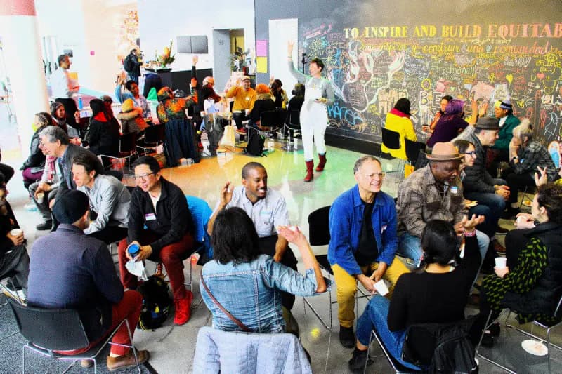 Interior gallery space filled with people mingling at what appears to be an opening reception or event, with yellow and black accents visible in the crowd.