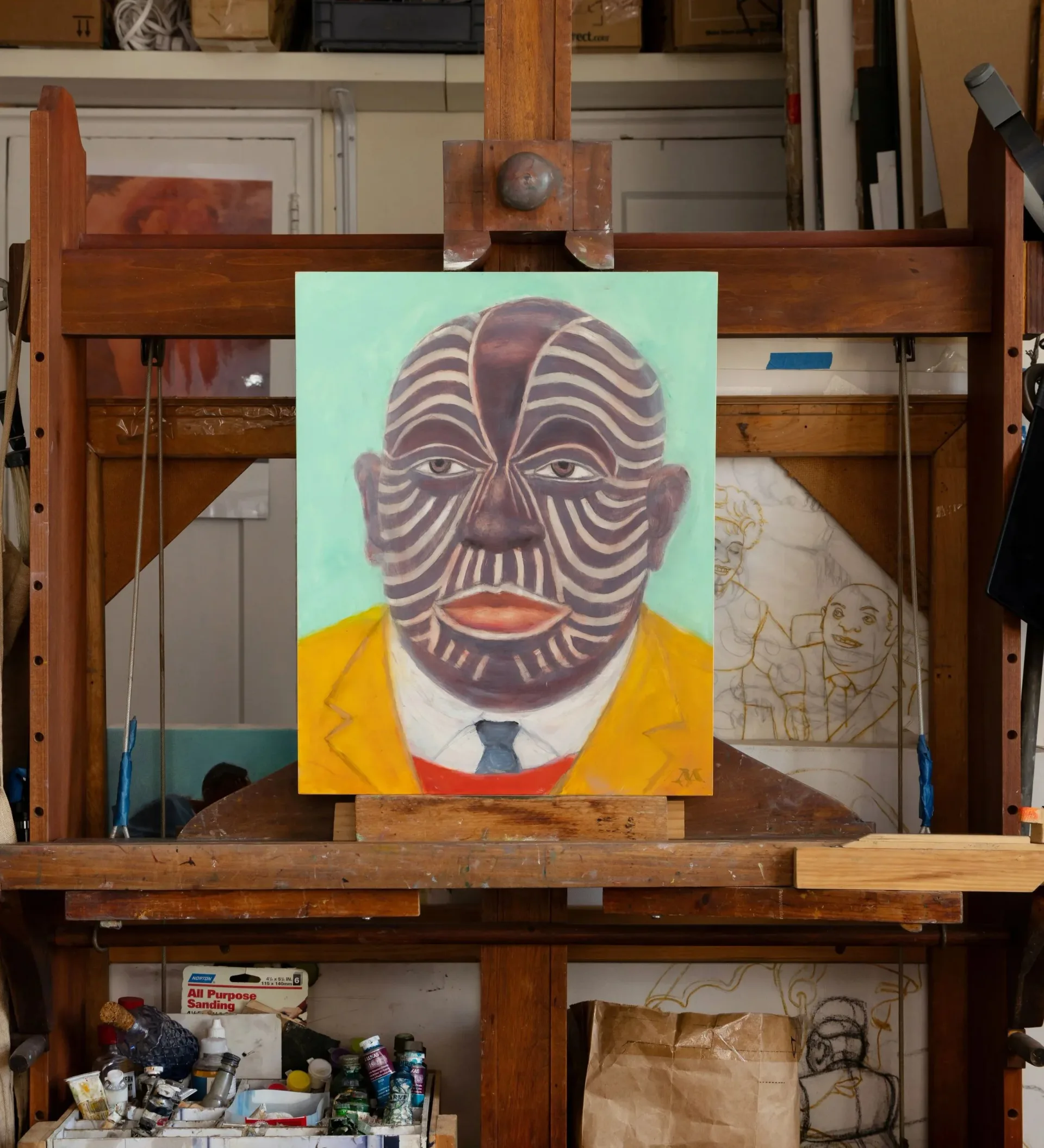 Studio workspace with wooden shelving unit displaying portrait painting with striped geometric face, yellow clothing, and sketches on surrounding surfaces.