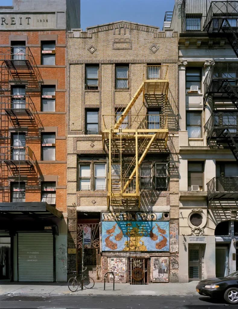 Photograph of a narrow brick building facade on a New York City street with yellow metal fire escape stairs, storefront with blue signage below, flanked by adjacent buildings.