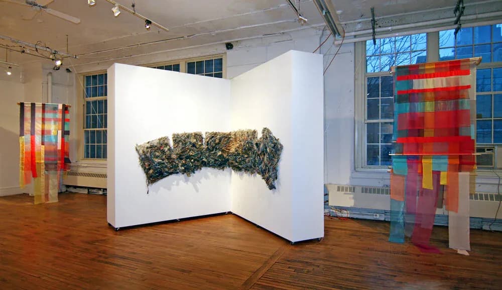 Gallery space with wooden floor displaying mixed-media wall installations in vibrant colors including red, yellow, and blue geometric patterns and organic forms.