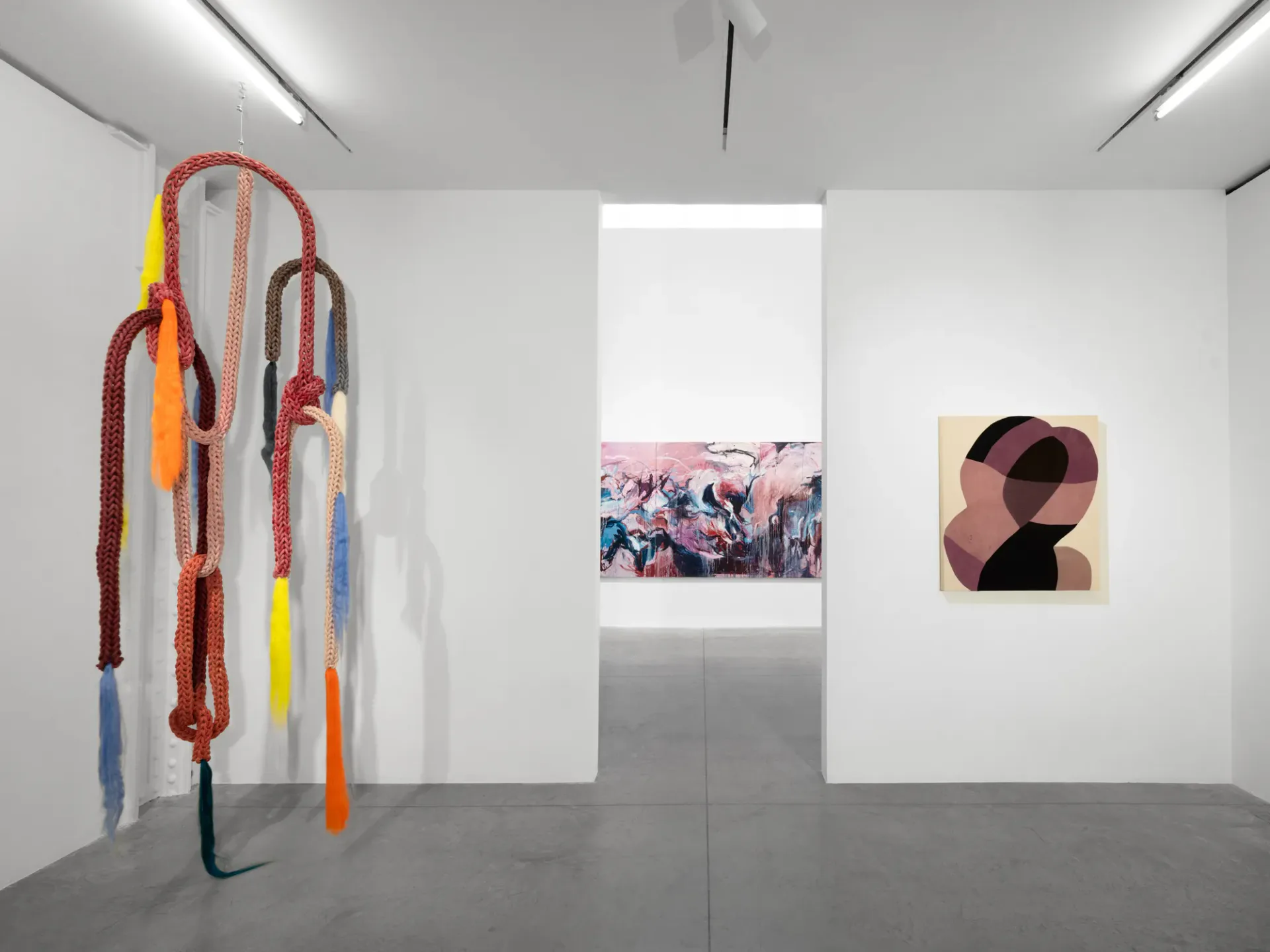 White gallery space with colorful rope coil installation on left, pink-toned photograph in center alcove, and abstract artwork on right.