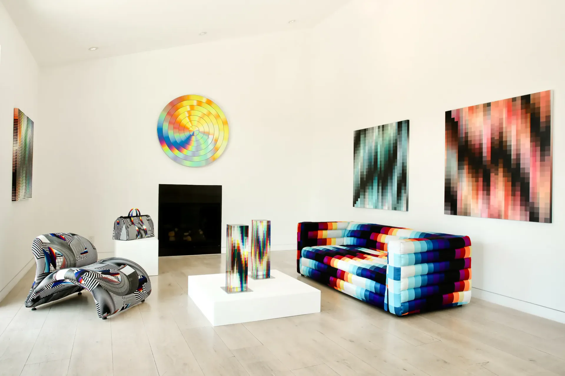 Gallery space with colorful geometric artworks including yellow spiral circle, pixelated abstract paintings, fabric sculptures, and metallic objects.