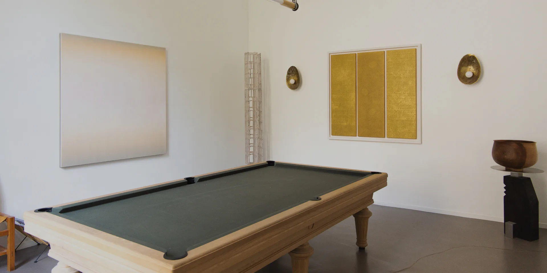 Gallery interior with pool table, white walls displaying abstract paintings and three-panel gold artwork, wooden and ceramic sculptural objects.