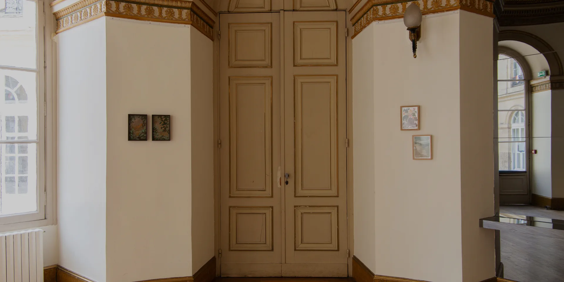 Classical gallery entrance with ornate wooden doors, white curved walls, small framed artworks, period architectural details.