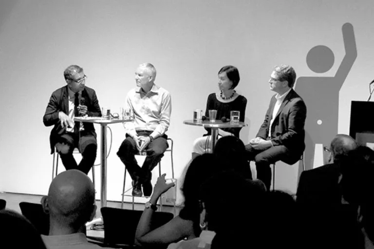 Panel discussion with four speakers seated in chairs on stage, audience visible in foreground, minimalist white backdrop with geometric graphics.