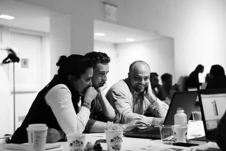 Three people sitting at a table with laptops in a bright white conference or workshop space, others visible in background.