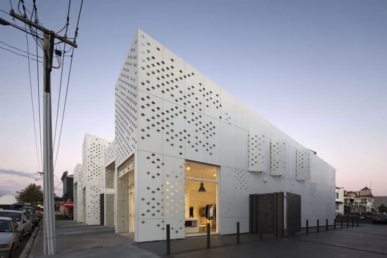 White contemporary building with perforated facade creating dot pattern, illuminated golden entrance, photographed at dusk with utility lines visible.