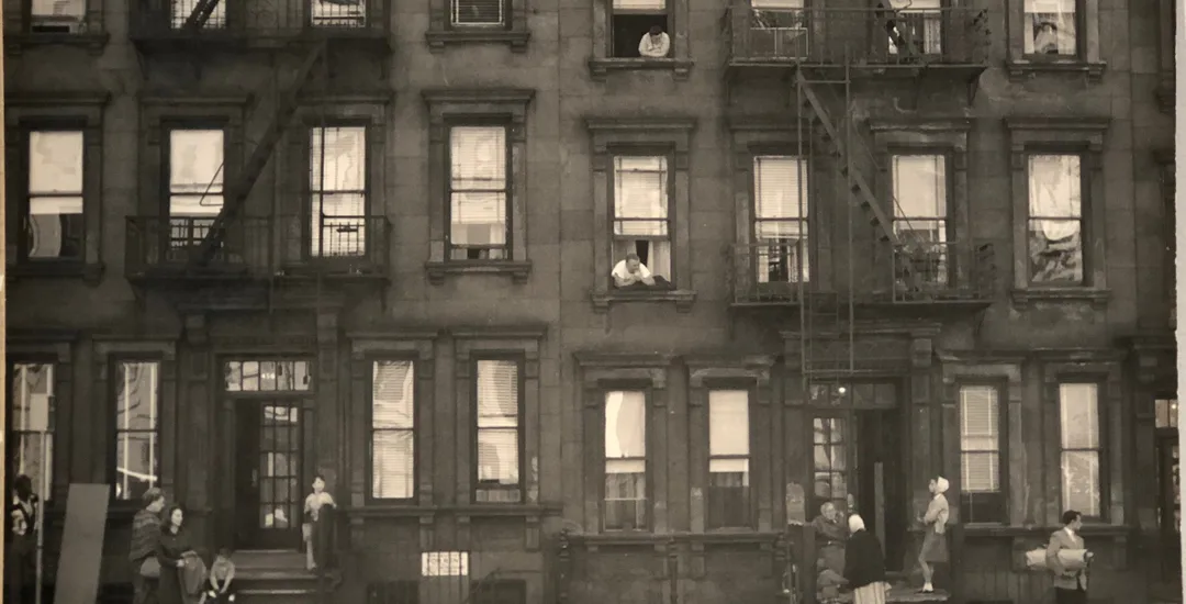 Photograph of a historic residential building facade with multiple rows of tall windows, stone masonry, and figures visible in some windows; appears to be a Chelsea townhouse or apartment building.