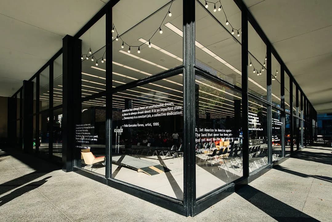 Interior view of a minimalist gallery space with black steel structural frames, glass partitions, white ceiling with track lighting, and polished concrete flooring.