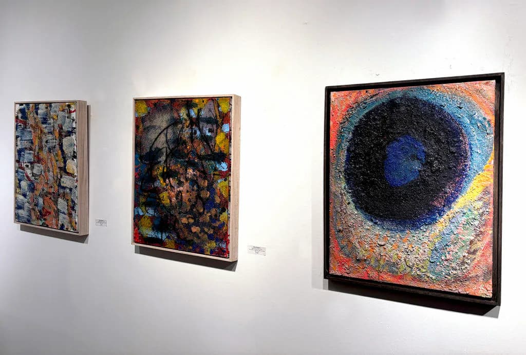 Gallery interior showing three framed abstract artworks on white wall, including textured paintings and a circular composition.
