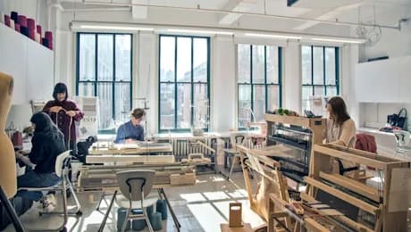 Bright studio workspace with large windows, wooden workbenches, and several people engaged in hands-on creative activities.
