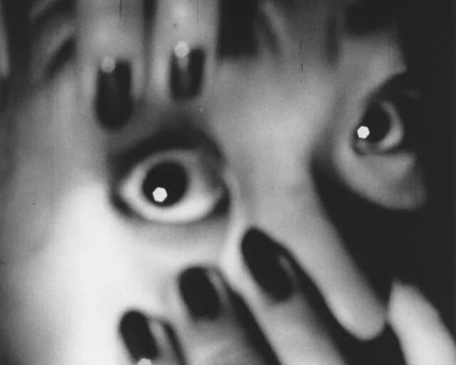 Black and white close-up photograph of a face with wide eyes and dark nail polish on raised hands, expressing shock or surprise.