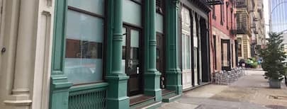 Storefront exterior with green-painted window frames and black doors on a urban street lined with buildings.
