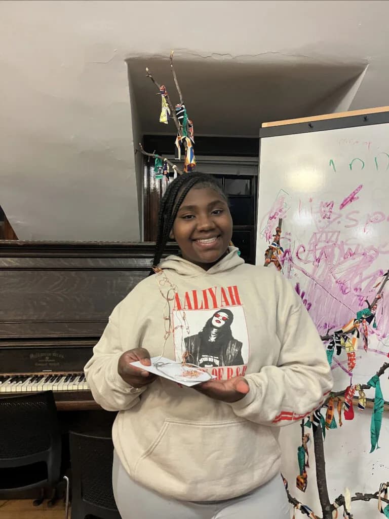 Young person in cream hoodie smiling in a studio space with piano, decorated whiteboard, and hanging artwork visible.