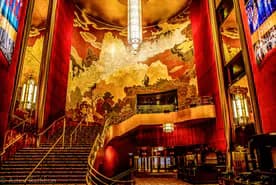 Interior of a grand Art Deco theater with ornate red and gold wall panels, a dramatic curved staircase, and elaborate sculptural ceiling details.