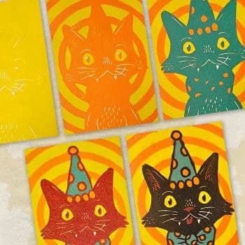 Colorful painted paper panels featuring stylized cat faces wearing party hats in orange, green, red, and black against bright yellow backgrounds.