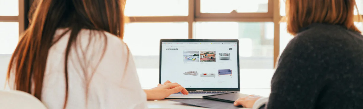 Two women viewed from behind sit at a table looking at a laptop screen displaying artwork thumbnails, near a bright window