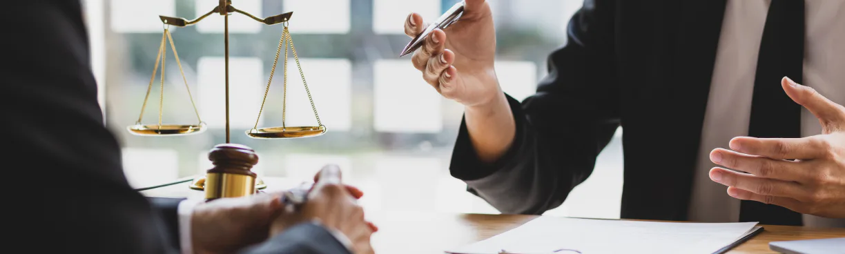 Close-up of a brass scale of justice and a gavel on a desk while a person in a suit hands over a pen