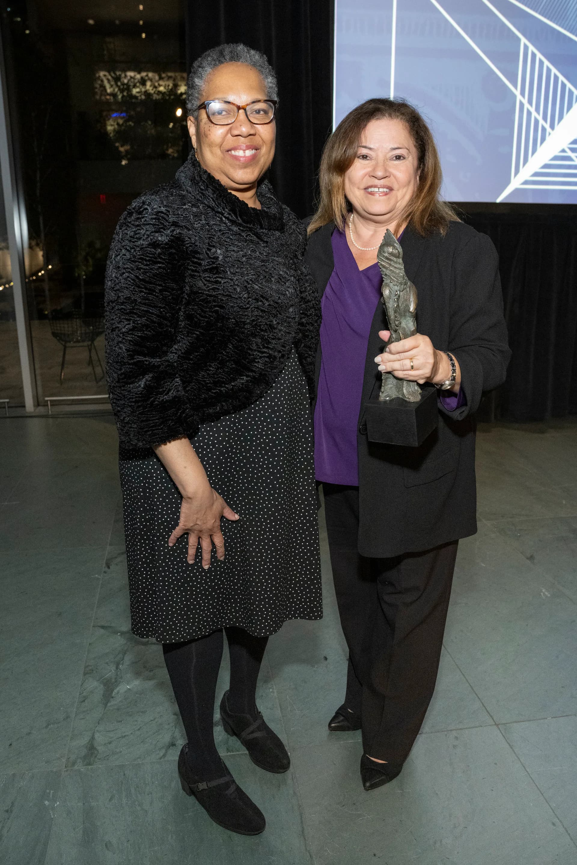 Two smiling women posing together indoors: one wearing glasses and a charcoal textured jacket, the other in a purple top and black blazer.