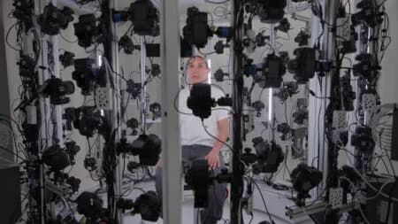 Man stands inside a photogrammetry rig surrounded by dozens of cameras and lights arranged in vertical rows, captured for an Art21 documentary segment.