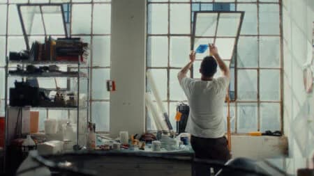 Artist in a white t-shirt reaches up to adjust a window pane in a bright studio filled with shelves of art supplies, seen from behind, an Art21 studio scene.