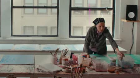 Artist in a headscarf leans over a table covered with paintbrushes, jars, and paint-splattered paper in a sunlit studio with large windows, an Art21 studio view.