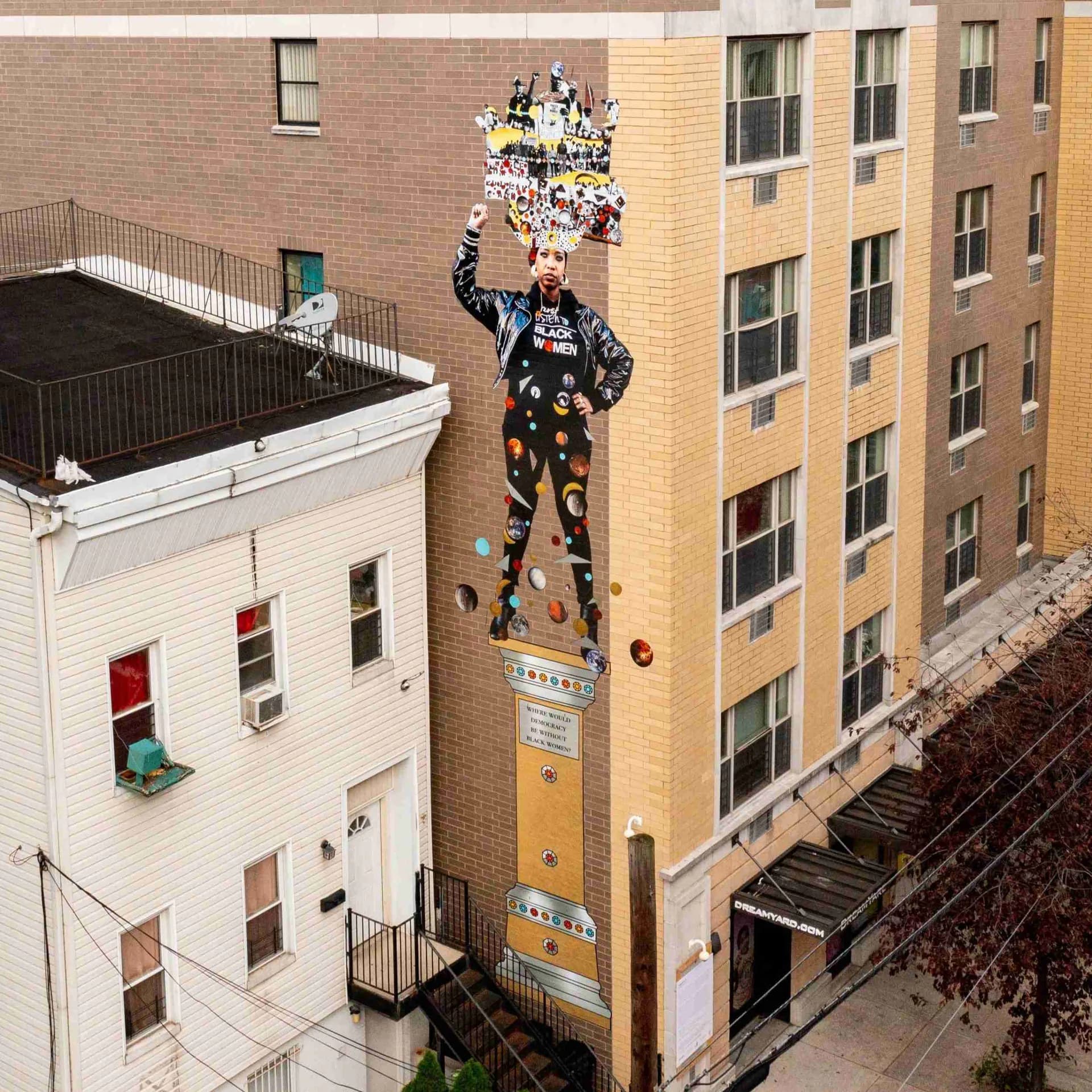 Tall mural on building facade featuring a collaged figure in dark clothing with multicolored abstract elements and floating spheres against a yellow vertical panel.