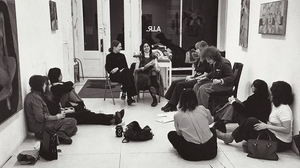 Black and white photograph of people seated in a circle on the floor of a white-walled gallery space during what appears to be a group discussion or critique session.