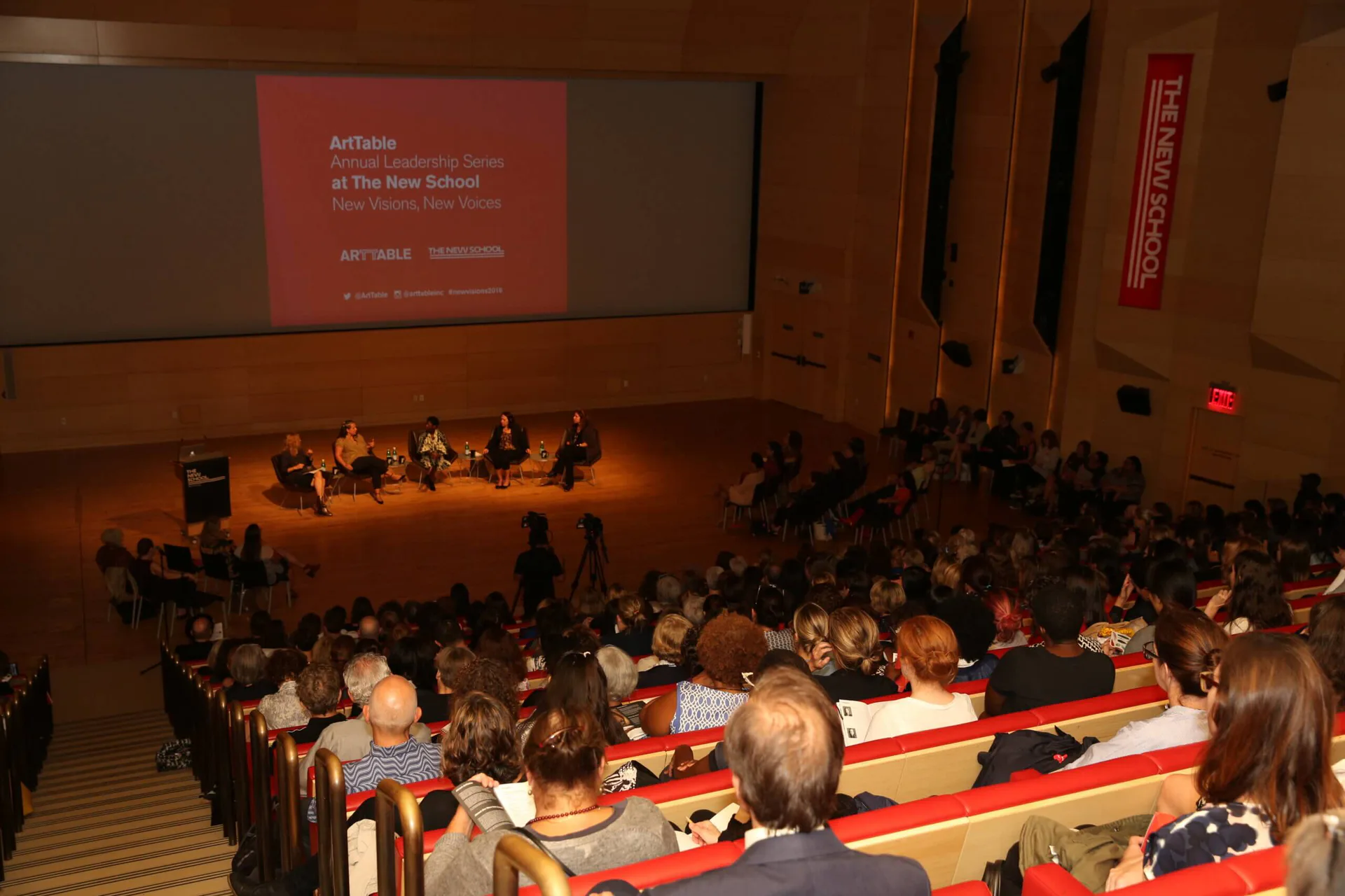 Interior of auditorium with audience seated facing stage; red panel visible on projection screen; warm lighting on wooden floor.