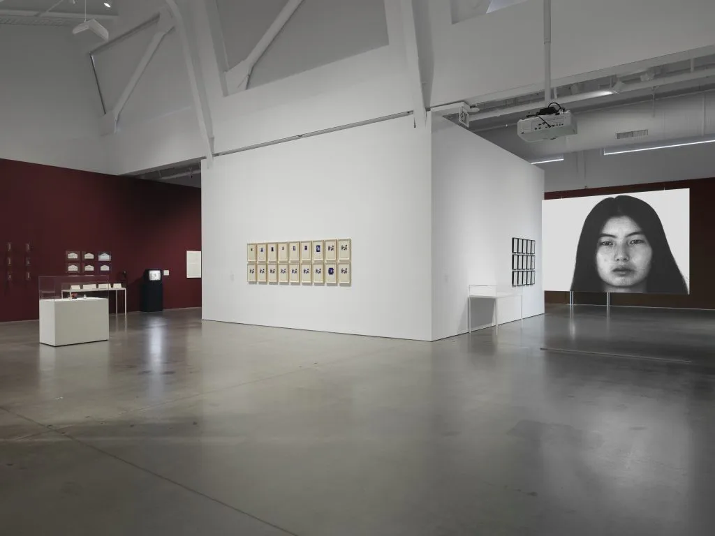 Gallery interior at Asia Art Archive in America showing white walls, polished concrete floor, burgundy accent wall, artworks on display, and projected video of a woman's face.
