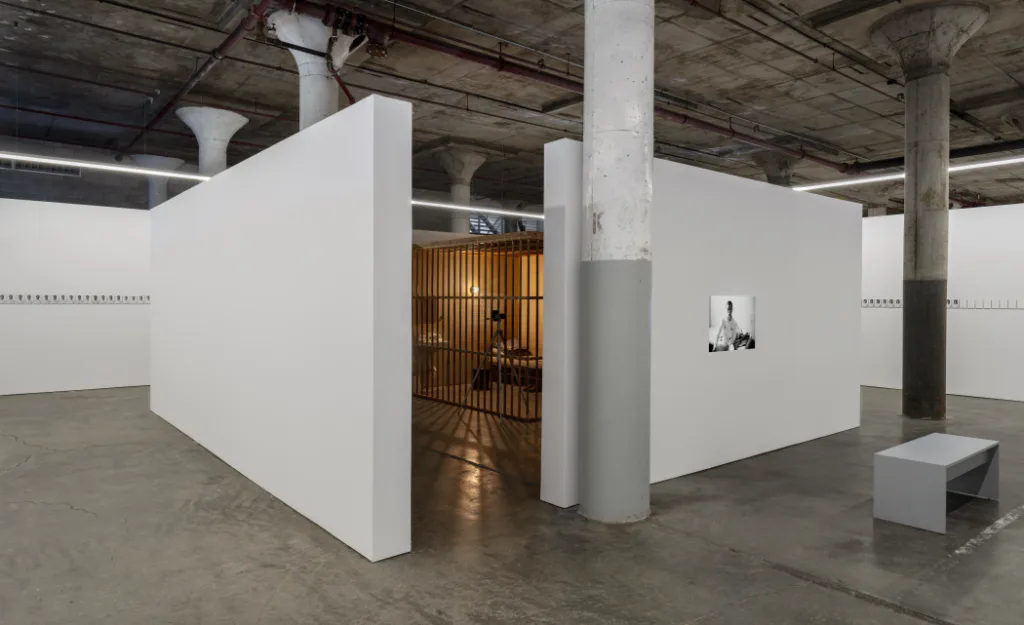 Gallery interior with white-walled rooms, industrial ceiling, concrete pillars, wooden warm-lit installation space visible through entryway.