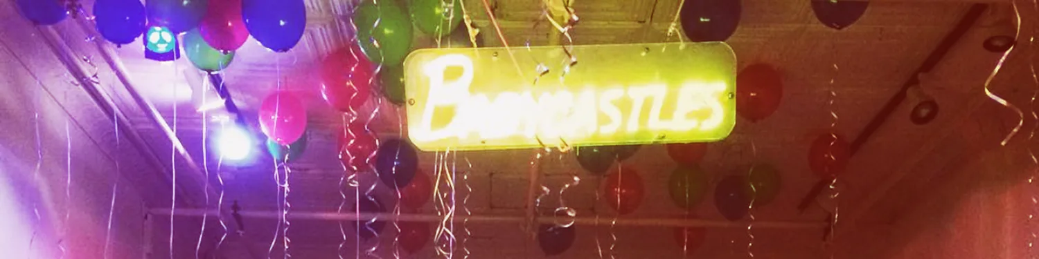 Neon sign reading "Babycastles" in yellow-green light, suspended amid colorful hanging spheres and tinsel against a dark ceiling.