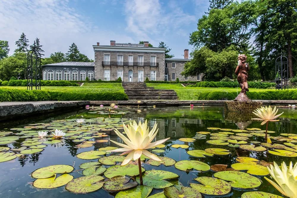 Exterior photograph of gray stone mansion with pond in foreground, yellow water lilies, lush landscaping, and reflected sky.