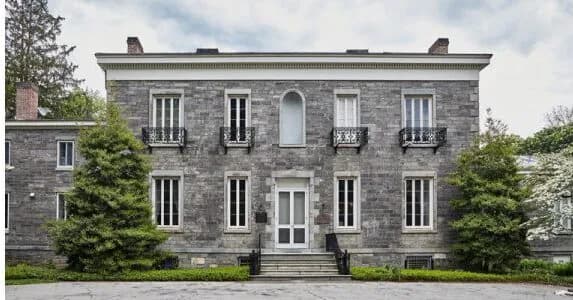 Exterior photograph of gray stone building with symmetrical windows, white door, ivy-covered walls, manicured hedges, and overcast sky.