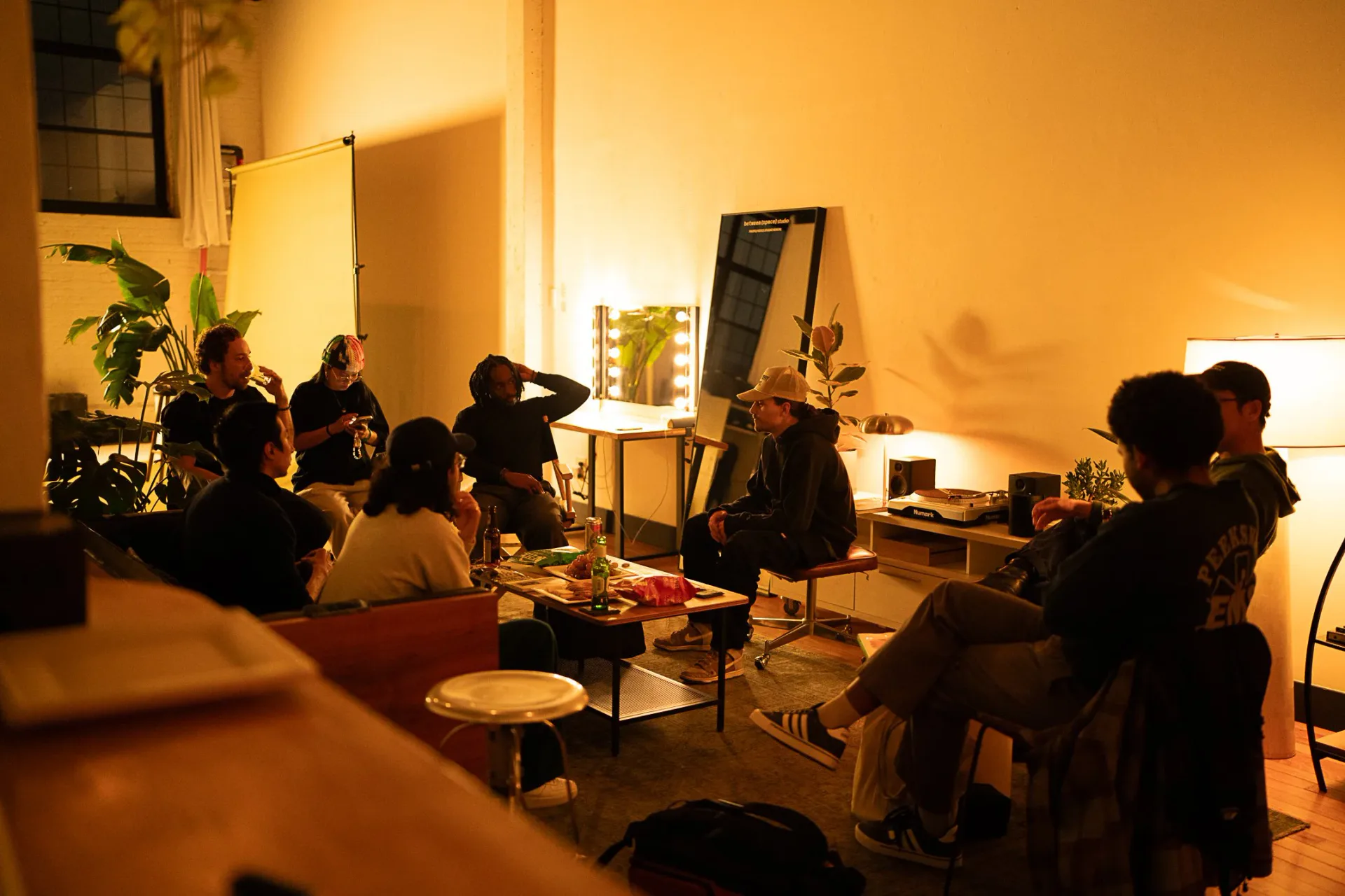 Group of people gathered in warmly lit interior space of Between Space Studio with yellow ambient lighting, plants, and minimal furniture.