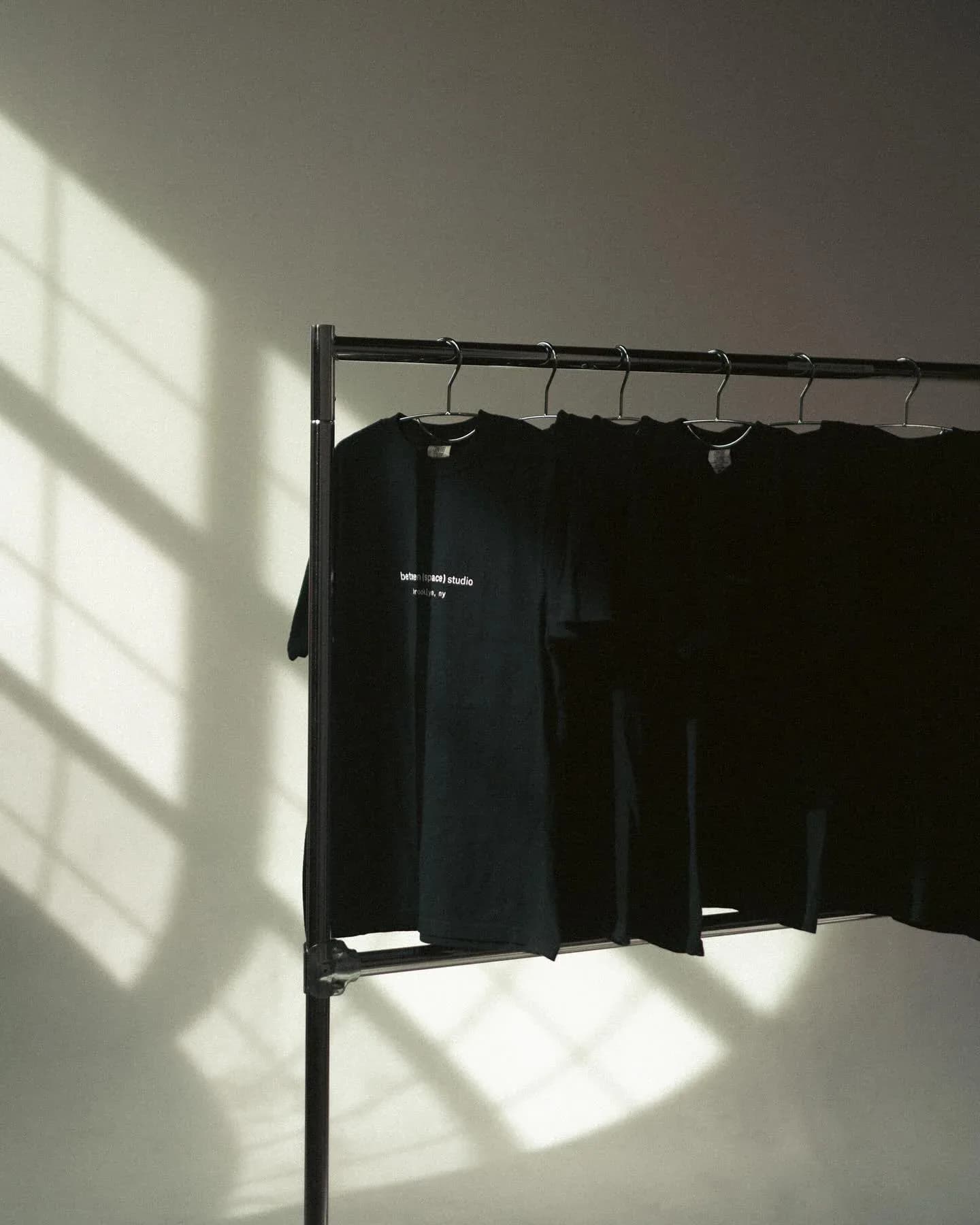 Black garments hanging on metal rack against white wall with bright window light casting rectangular shadows.