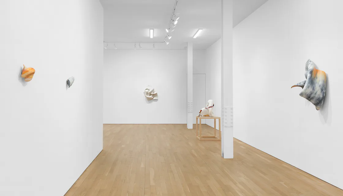 White gallery interior with wooden floor, minimal artwork mounted on walls, track lighting, and sculptural pieces on pedestals.