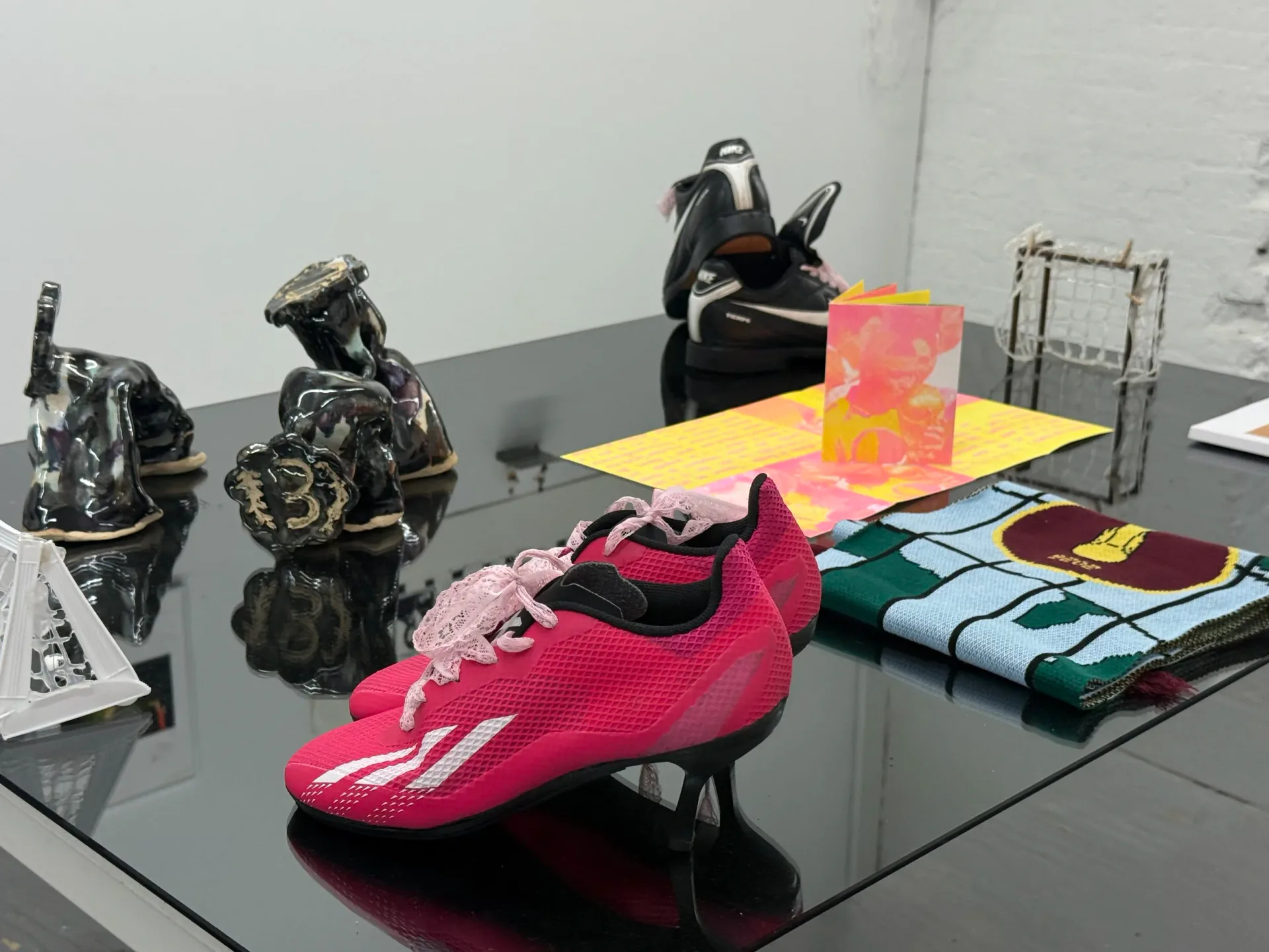 Gallery table displaying metallic sculptural objects, pink and coral textiles, a bright pink shoe, and folded fabric swatches in various colors.