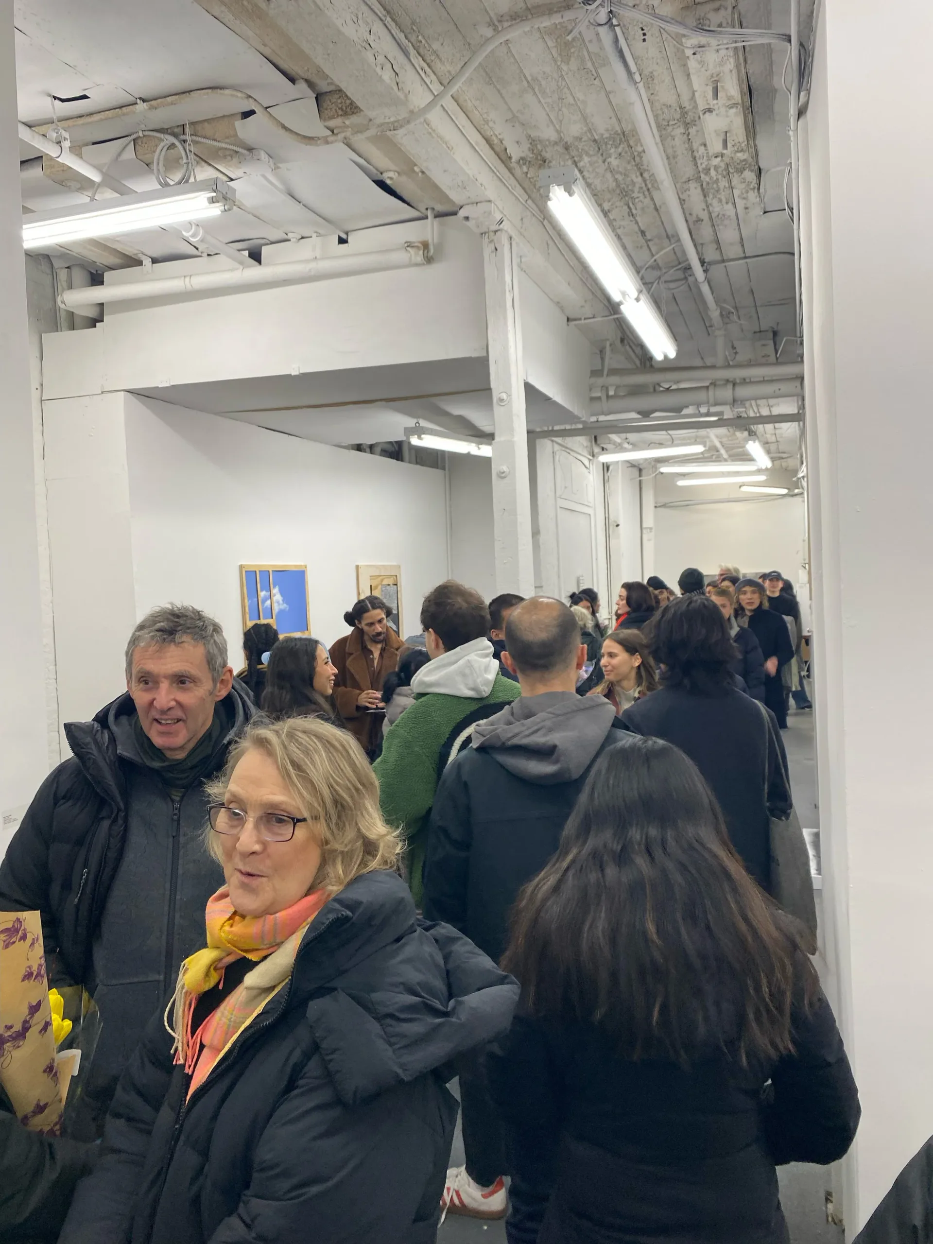 Gallery interior with industrial ceiling and visitors gathered; small blue artwork visible on wall in background.