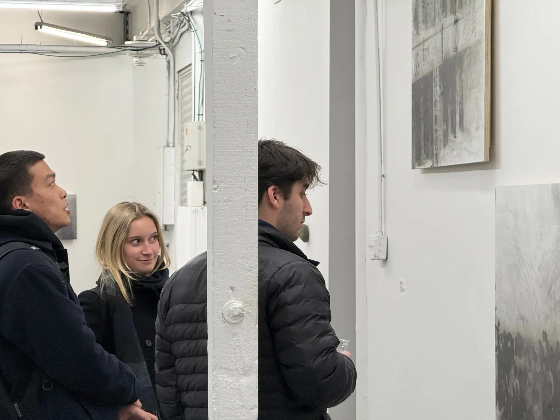 Three visitors in dark jackets viewing large framed gray-scale artwork on white gallery walls.