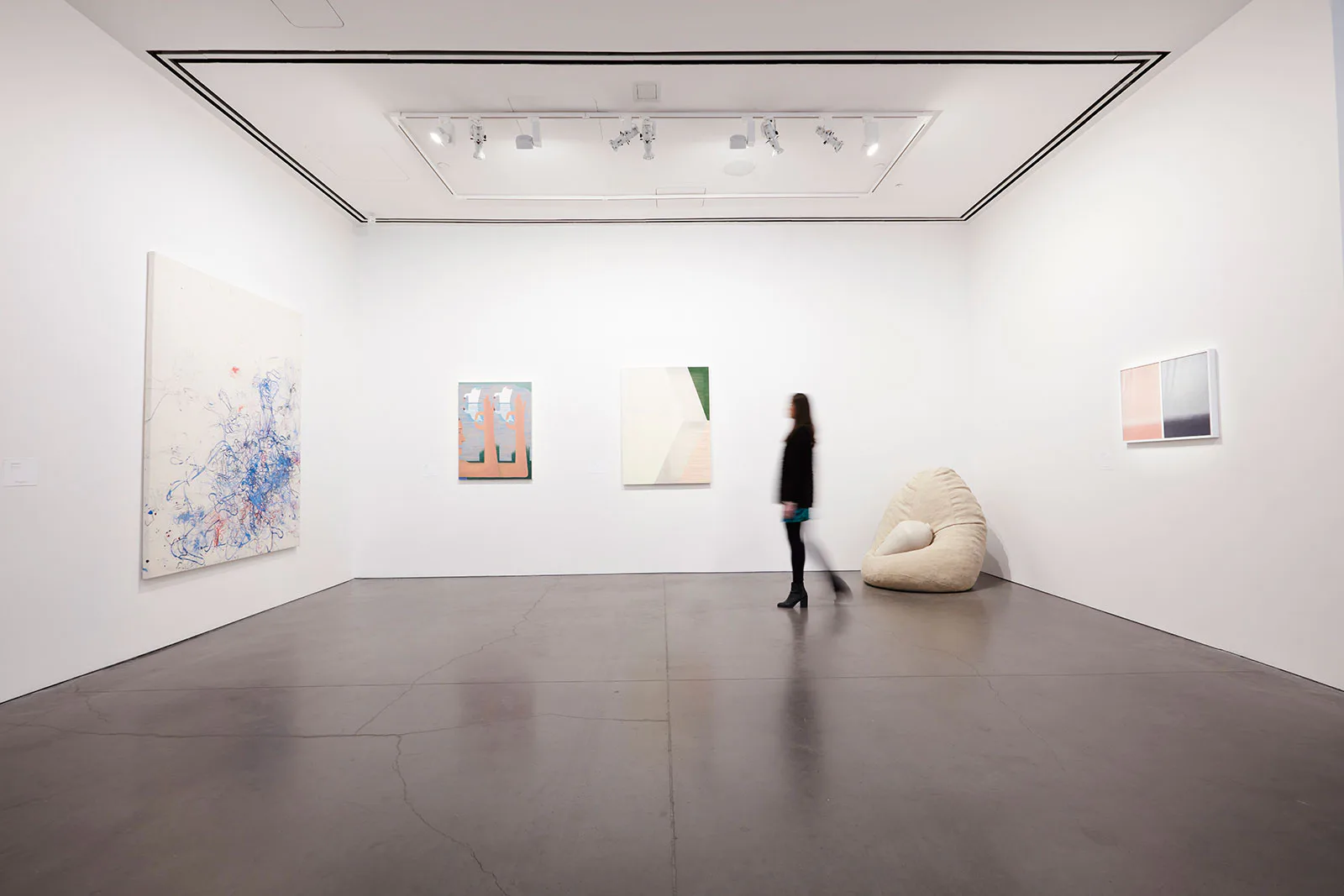 Gallery interior with white walls, track lighting, several artworks, a cream bean bag chair, and a woman walking across the concrete floor