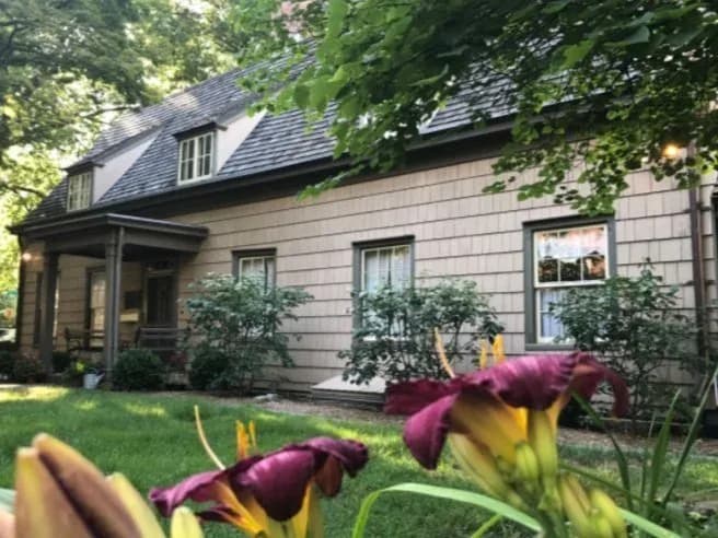 Exterior of the Bowne House, a historic shingled colonial-style house with dormer windows and a covered porch, surrounded by greenery and flowers