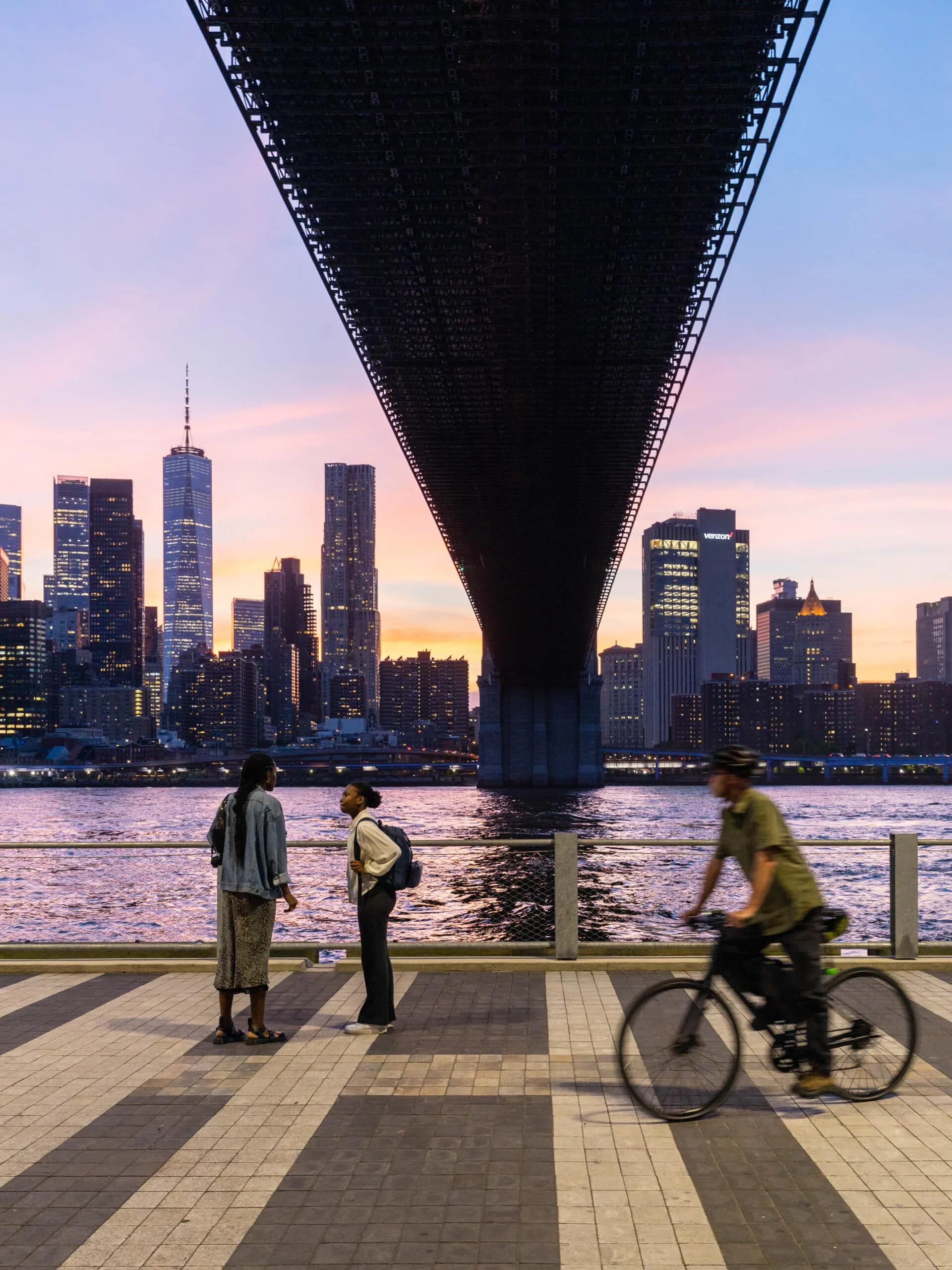 Photograph taken at dusk showing Manhattan skyline framed beneath a massive bridge underside, with pedestrians and cyclist on a waterfront promenade during golden hour.