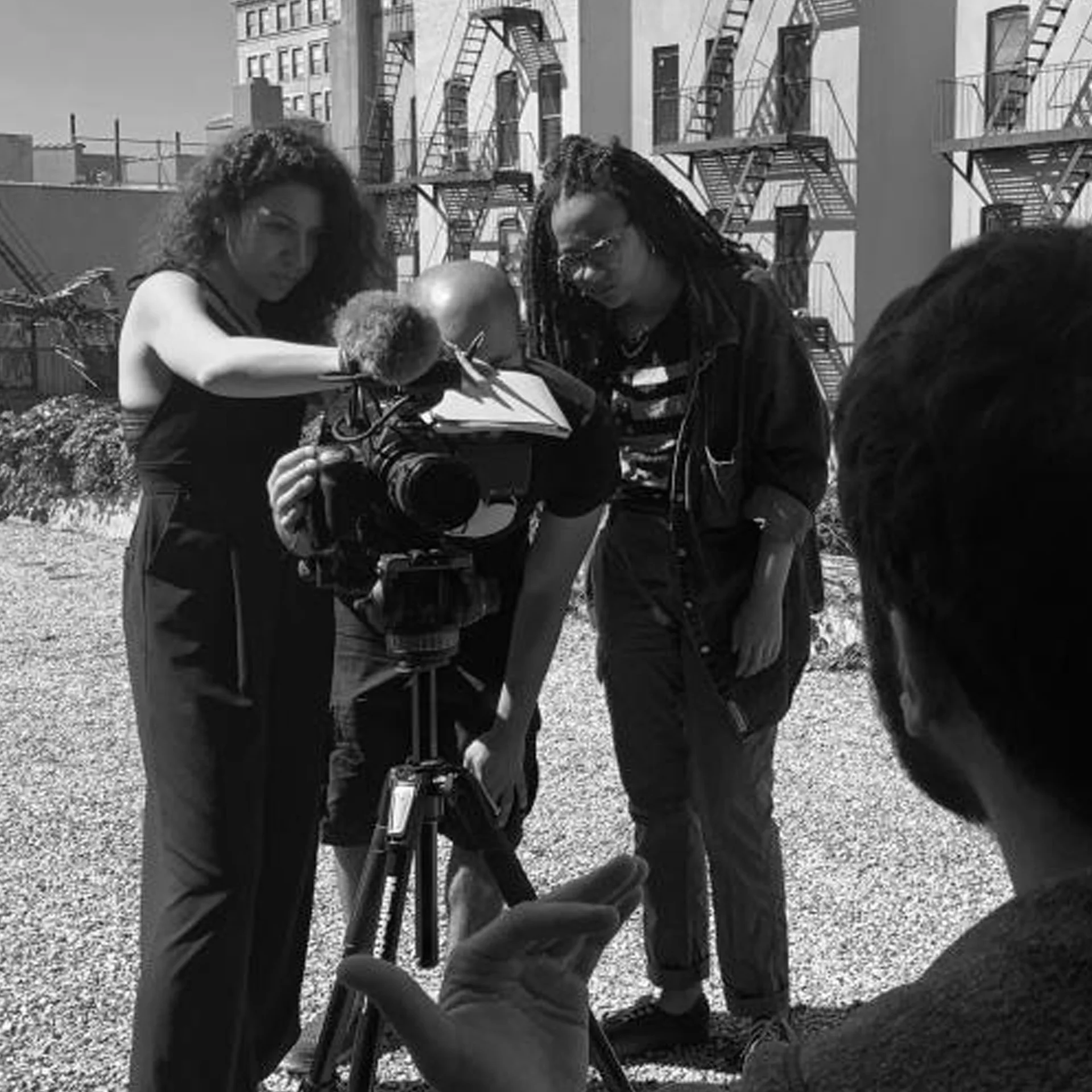 Black and white photo of three people setting up a video camera on a tripod outdoors near fire escapes, likely a Bronx Documentary Center production shoot
