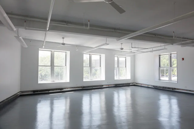 Empty gallery room with white walls, exposed ceiling pipes, tall windows, and a gray reflective floor