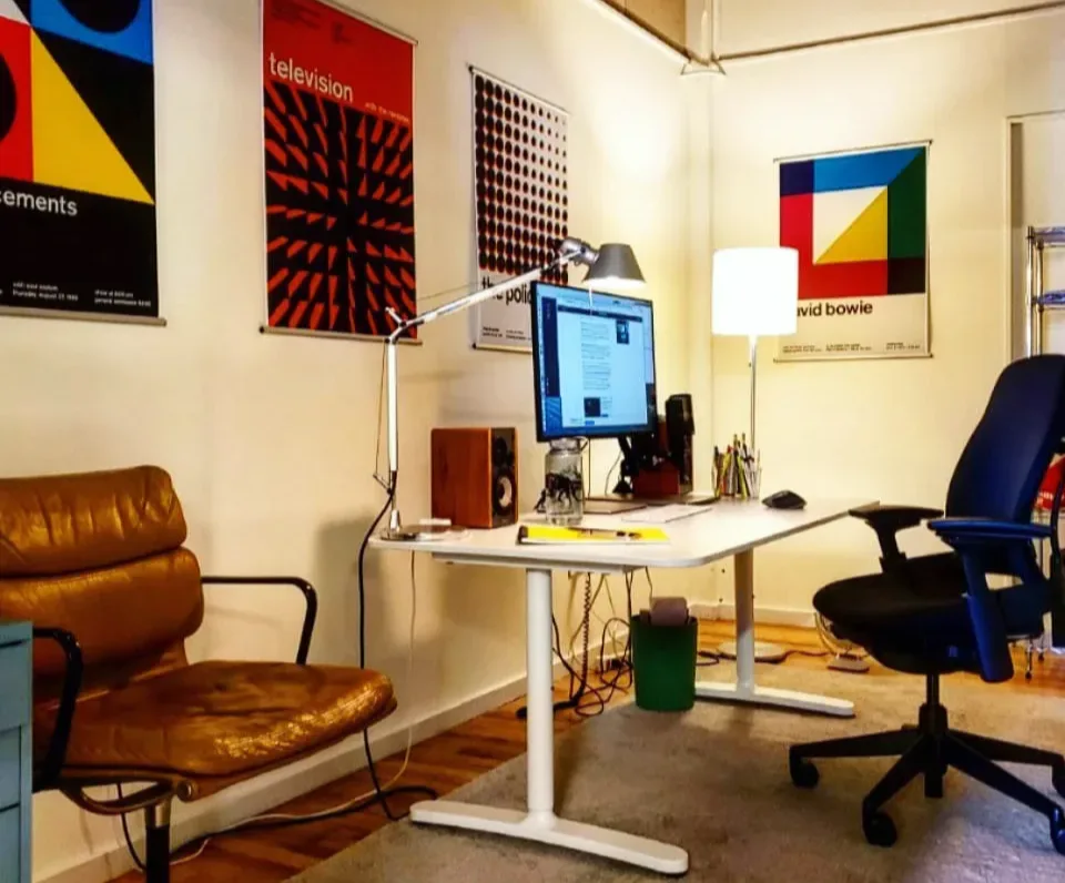 Studio office space with desk, computer monitor, brown leather chair, blue office chair, and colorful abstract posters on walls.