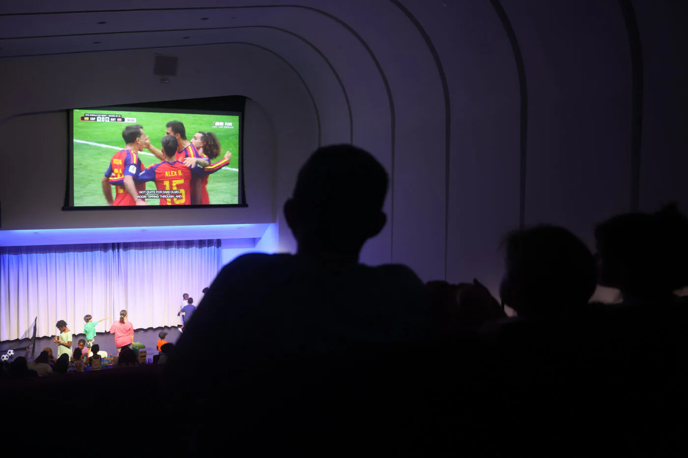 Interior of Brooklyn Children's Museum with silhouetted visitors watching a large screen displaying children's content, blue accent lighting on white architectural elements.